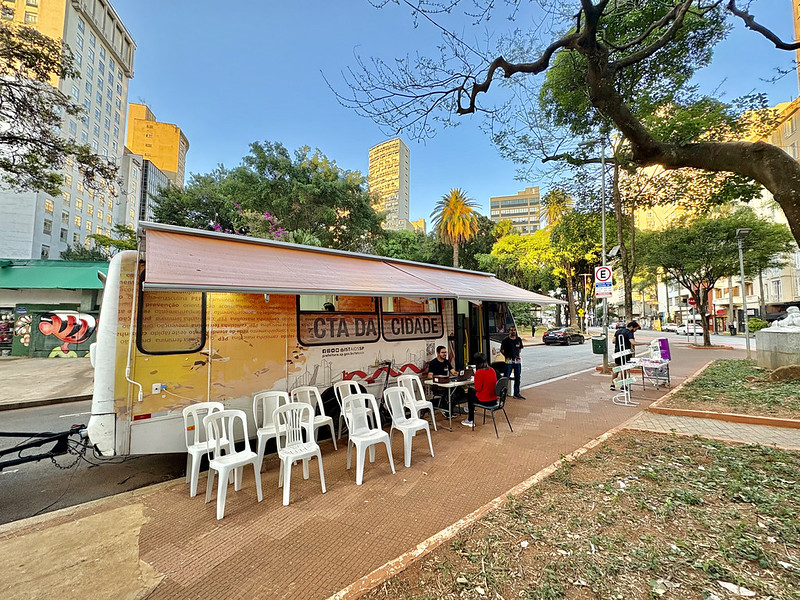 A imagem mostra uma unidade móvel de atendimento instalada em uma praça ou área pública arborizada de uma região central da cidade.  Em destaque está um trailer adaptado, identificado como “CTA da Cidade”, sigla normalmente associada aos Centros de Testagem e Aconselhamento em saúde. O veículo possui uma cobertura retrátil aberta sobre a calçada, criando uma área de atendimento ao público. Em frente ao trailer há várias cadeiras plásticas brancas organizadas para espera, enquanto algumas pessoas conversam ou realizam atendimentos em mesas instaladas ao lado da unidade.  O cenário urbano é composto por prédios altos ao fundo, árvores de grande porte, palmeiras e uma praça com áreas de circulação de pedestres. A luz do sol ilumina a cena, sugerindo que a atividade ocorre durante o dia.