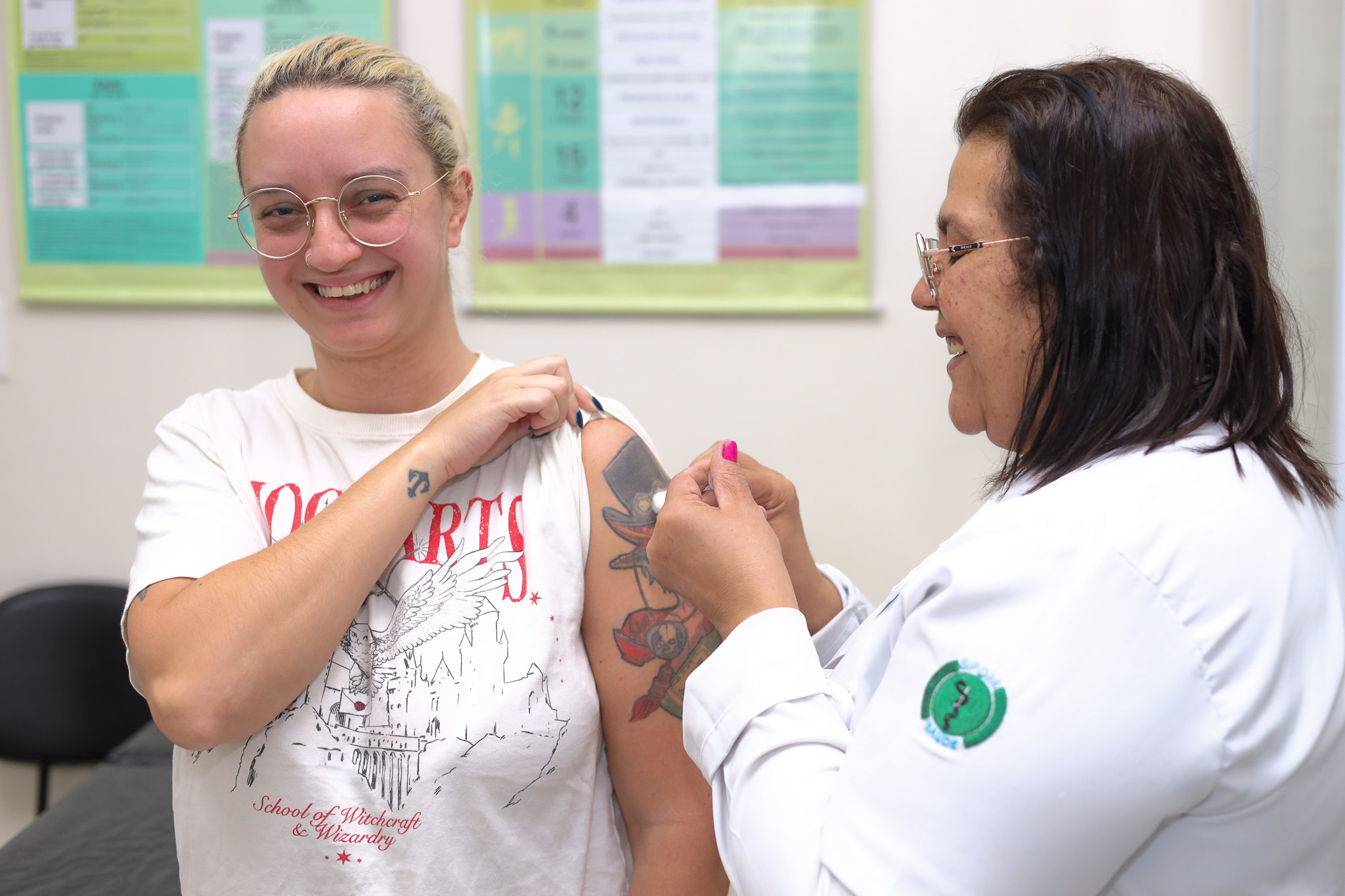 Em uma sala de atendimento de saúde, uma mulher jovem de cabelos loiros presos, usando óculos e camiseta branca, sorri enquanto recebe uma vacina no braço esquerdo. Ela levanta a manga da camiseta para facilitar a aplicação. Ao lado, uma profissional de saúde com jaleco branco e óculos aplica a injeção com uma seringa, concentrada no procedimento. No fundo da sala há cartazes informativos coloridos fixados na parede, típicos de ambientes de unidades de saúde. A cena transmite um momento de cuidado e prevenção em saúde.