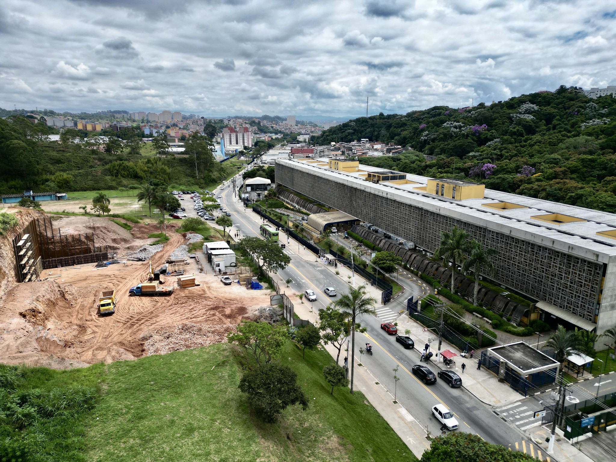 A imagem mostra uma vista aérea da região de Cidade Tiradentes, na zona leste de São Paulo. Em primeiro plano, há um canteiro de obras com movimentação de terra, máquinas e caminhões, indicando a construção de um novo equipamento público — possivelmente uma unidade de saúde. Ao lado, uma avenida com fluxo de carros e ônibus conecta o bairro. À direita, destaca-se um grande edifício institucional já existente, cercado por área verde e relevo com morros e vegetação. Ao fundo, vê-se a paisagem urbana com conjuntos habitacionais característicos da região, sob um céu nublado.