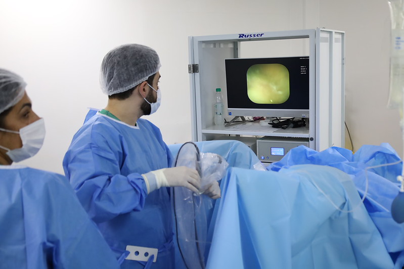 A imagem mostra um procedimento médico realizado em ambiente hospitalar ou centro cirúrgico.  Em primeiro plano, aparecem profissionais de saúde usando aventais cirúrgicos azuis, máscaras, luvas e toucas descartáveis, seguindo protocolos de assepsia. Um dos profissionais segura um equipamento flexível semelhante a um endoscópio enquanto observa atentamente um monitor.  Na tela é exibida uma imagem interna do corpo humano captada por vídeo, indicando que se trata de um procedimento minimamente invasivo, como uma endoscopia, colonoscopia, broncoscopia ou outro exame/intervenção guiado por câmera. O paciente está coberto por campos cirúrgicos azuis estéreis, que ocupam boa parte da cena.  O ambiente é limpo, bem iluminado e equipado com tecnologia médica. A fotografia transmite uma atmosfera de cuidado especializado, precisão técnica e trabalho em equipe, destacando o uso de recursos diagnósticos e terapêuticos modernos na assistência à saúde.