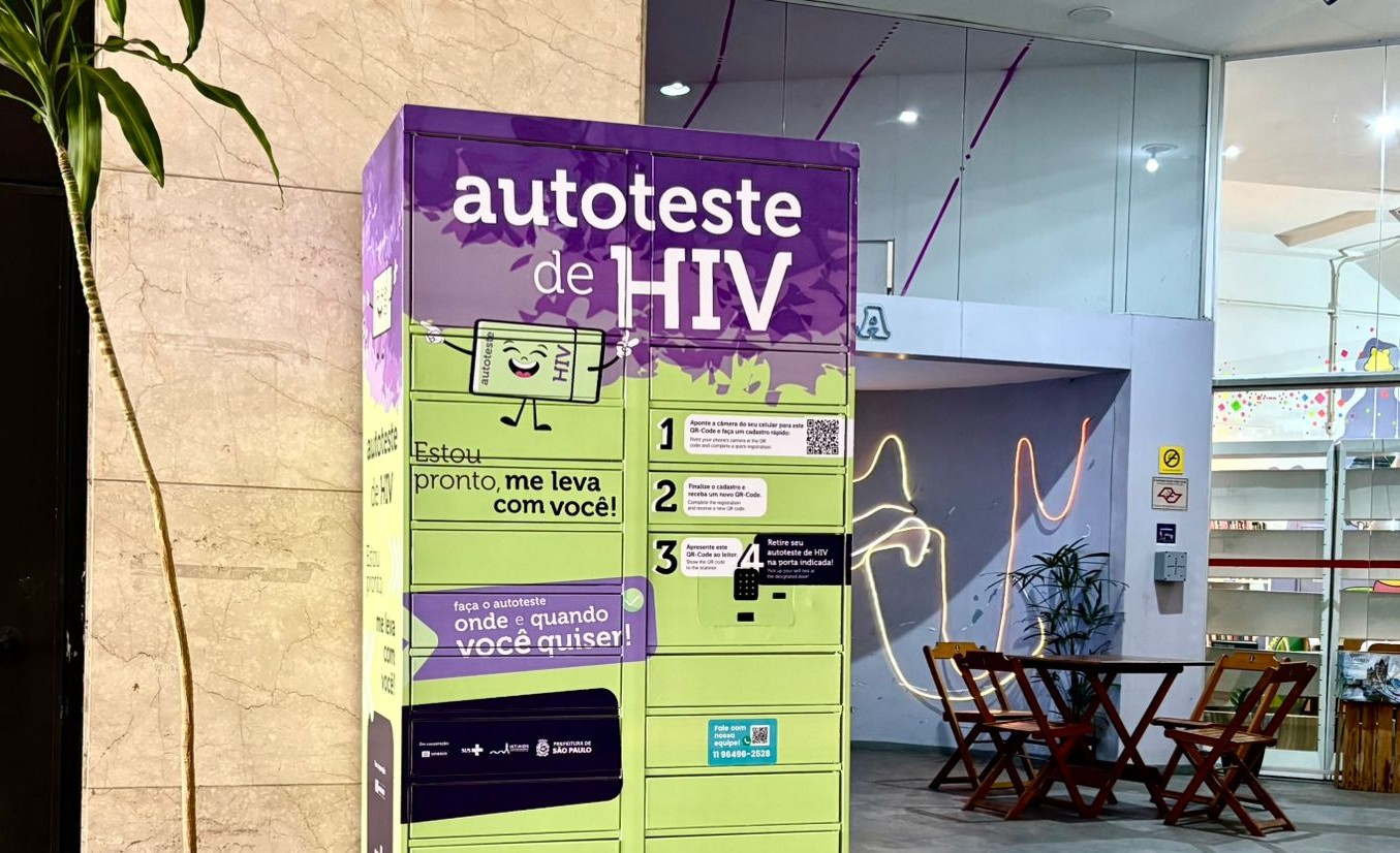 A imagem mostra uma máquina de distribuição de autotestes de HIV instalada em um corredor interno. O equipamento é colorido, com tons de roxo, verde e laranja, e exibe mensagens como “autoteste de HIV”, “Estou pronto, me leva com você!” e instruções ilustradas sobre como retirar e utilizar o teste. Há ícones e setas chamando a atenção para o serviço, além de logotipos de instituições públicas. Ao fundo, é possível ver um espaço interno com iluminação clara, paredes brancas, colunas e uma área que parece ser um ambiente de atendimento ou exposição, com prateleiras e itens coloridos. Uma planta alta aparece à esquerda da máquina.