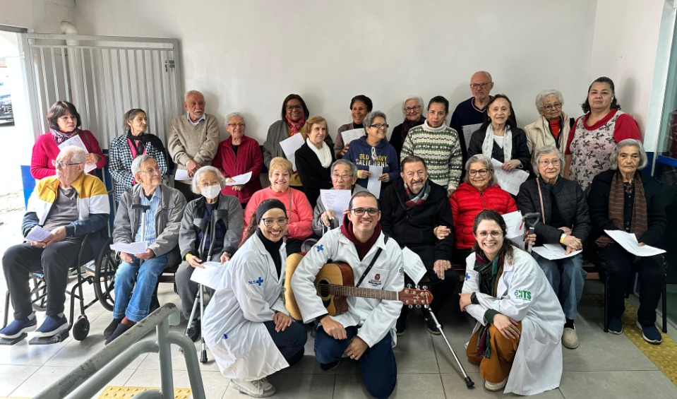 A imagem mostra um grupo grande de pessoas idosas reunidas em um espaço interno, aparentando participar de uma atividade coletiva. Elas estão sentadas e em pé, muitas segurando folhas de papel, como se estivessem envolvidas em uma dinâmica de canto, leitura ou grupo terapêutico. Na frente do grupo, três profissionais de saúde vestindo jalecos brancos posam sorrindo; um deles segura um violão, sugerindo que a atividade tem caráter musical ou recreativo. Ao redor deles, os idosos também sorriem e parecem engajados. Há cadeiras e uma cadeira de rodas entre os participantes, indicando acessibilidade e inclusão. O ambiente é simples e iluminado, com clima acolhedor e de convivência comunitária