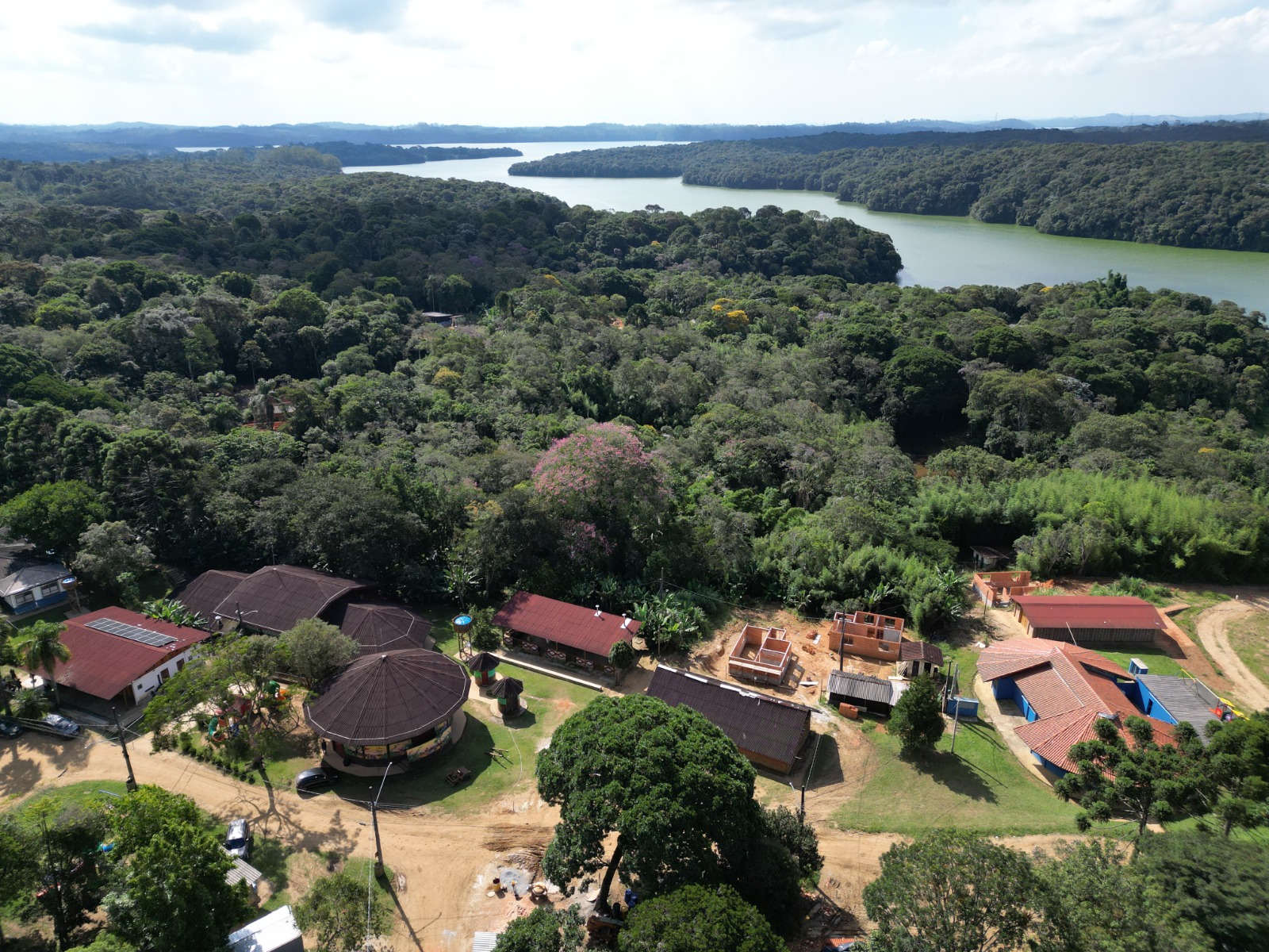 A imagem mostra uma vista aérea de uma área rural cercada por mata densa e um grande corpo d’água ao fundo, provavelmente uma represa ou rio de grande extensão. A paisagem é predominantemente verde, com vegetação abundante e contínua.  No primeiro plano, há um pequeno núcleo de construções distribuídas ao longo de uma estrada de terra. As edificações têm características simples, com telhados de barro e algumas estruturas em construção. Um dos destaques é um prédio circular com cobertura ampla, que sugere um espaço comunitário ou de convivência.  As casas e construções estão integradas ao ambiente natural, com árvores ao redor e poucos sinais de urbanização densa. A cena transmite a ideia de uma comunidade isolada ou de difícil acesso, inserida em uma área de preservação ambiental ou zona rural.