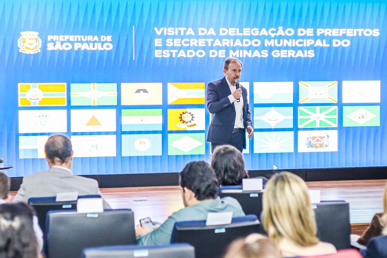 A imagem mostra um auditório durante uma apresentação institucional da Prefeitura de São Paulo. Em primeiro plano, aparecem participantes sentados, voltados para o palco, acompanhando a exposição. No centro da cena, um homem de terno segura um microfone e faz uma apresentação diante de um grande painel de LED. Ao fundo, o telão exibe a identidade visual da Prefeitura de São Paulo e a mensagem: