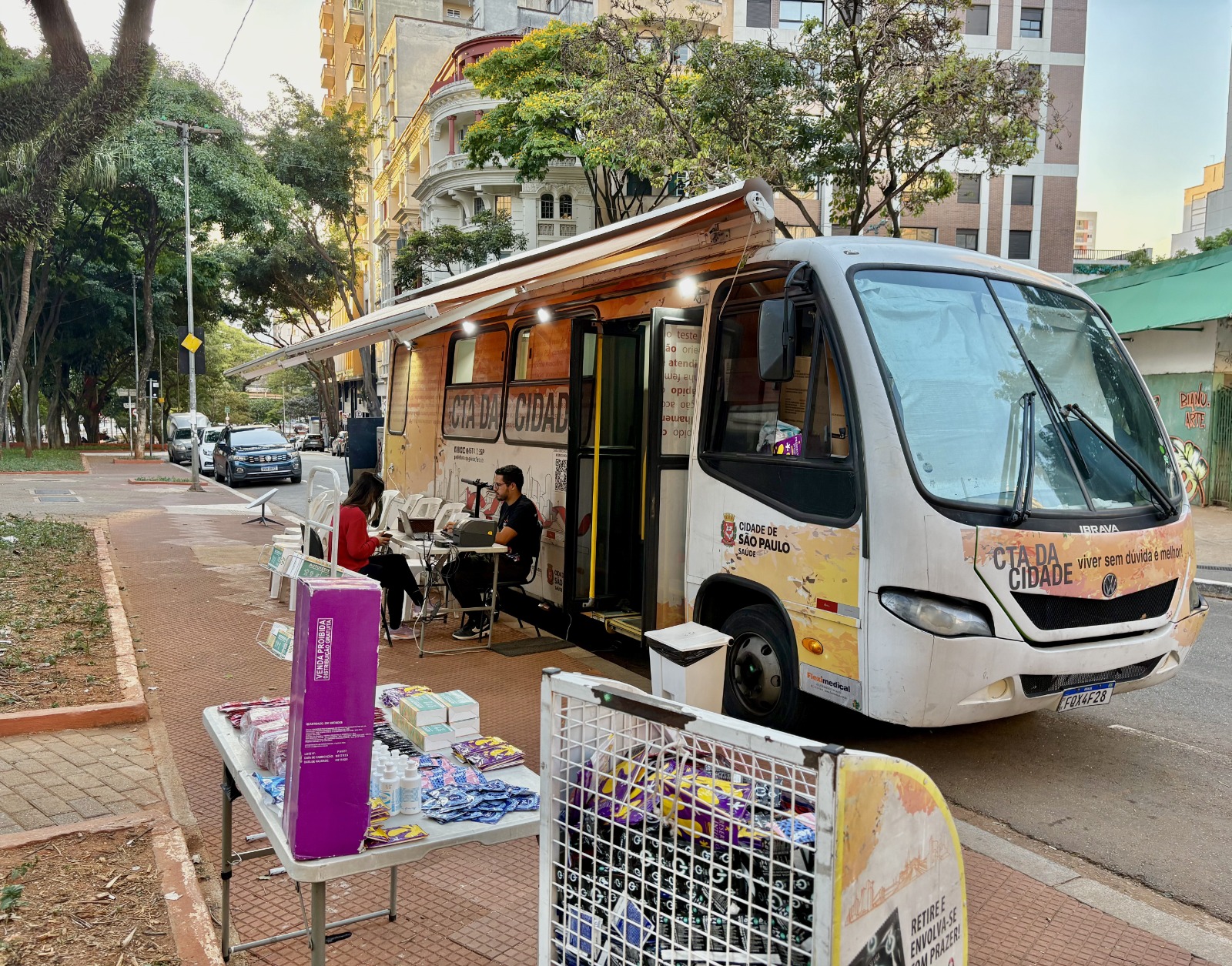 A imagem mostra uma unidade móvel de saúde estacionada em uma via urbana arborizada, aparentemente em uma praça ou avenida de São Paulo. O veículo é um ônibus adaptado do programa CTA da Cidade, da Prefeitura de São Paulo, identificado com a mensagem