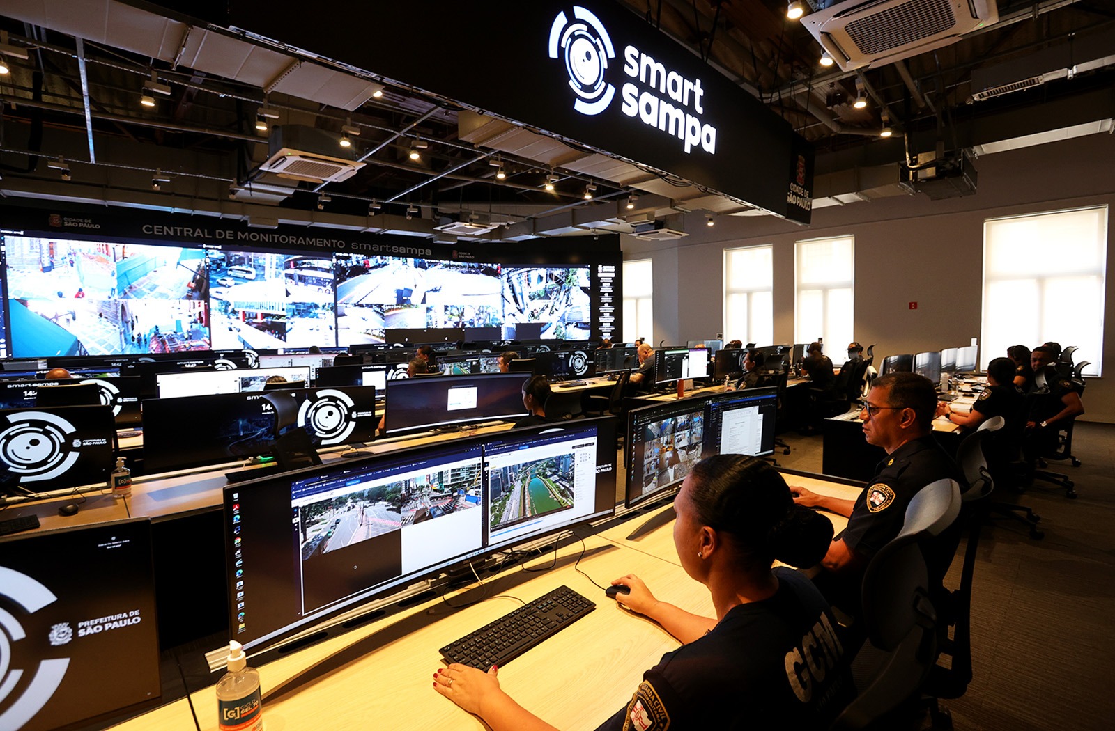 Sala ampla e moderna da Central de Monitoramento Smart Sampa, em São Paulo. No alto, o letreiro “Smart Sampa”. Ao fundo, um grande videowall mostra dezenas de câmeras das ruas da cidade em tempo real.  Em várias mesas, agentes da Guarda Civil Metropolitana (GCM) acompanham as imagens em múltiplos monitores. Em primeiro plano, uma mulher e um homem observam as telas. O ambiente é organizado, iluminado e operacional