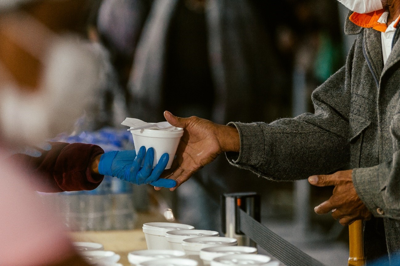Orientador social usando luva azul entrega um pote com sopa para um atendido da OBT. O munícipe usa um agasalho cinza.