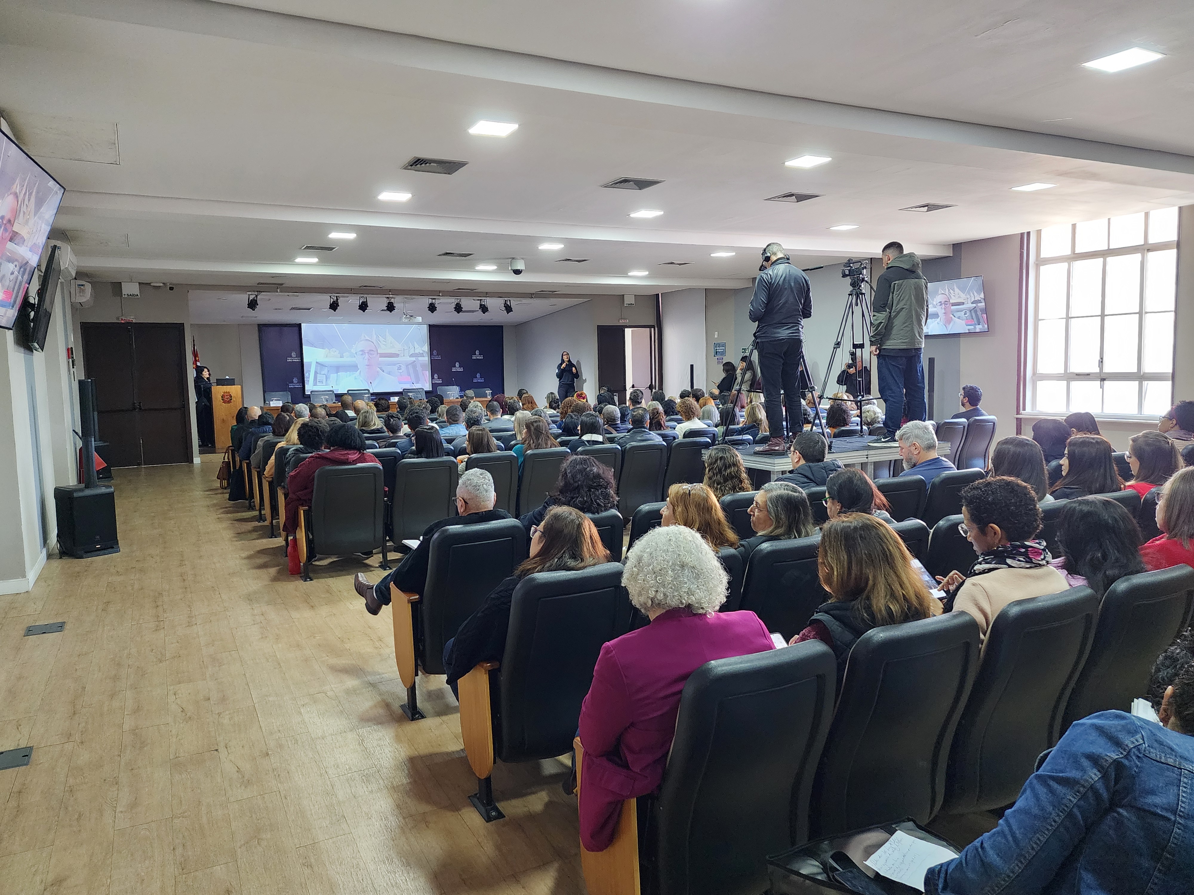 Imagem de fundo do auditório onde ocorreu o evento. As pessoas presentes na imagem estão de costas, sentadas em cadeiras dispostas em fileiras, atentos a palestra que está sendo transmitida no telão a frente.