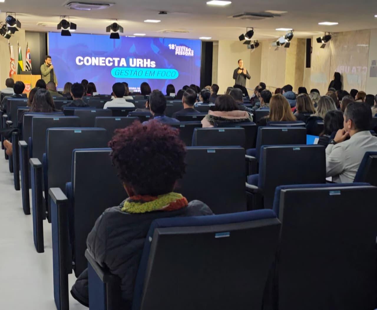 #PraTodosVerem: Fotografia de um auditório com dezenas de pessoas sentadas em cadeiras azuis, voltadas para um palco onde acontece uma apresentação. Ao fundo, um telão exibe a identidade visual do evento com os dizeres “Conecta URHs – Gestão em Foco” e a marca da 18ª Semana de Gestão de Pessoas. À esquerda do palco, um palestrante fala ao público com um microfone, enquanto, à direita, um intérprete de Libras realiza a tradução simultânea. O ambiente é iluminado, possui equipamentos de iluminação suspensos no teto e bandeiras do Brasil, do Estado de São Paulo e do Município de São Paulo ao lado do palco. A plateia acompanha atentamente a apresentação.