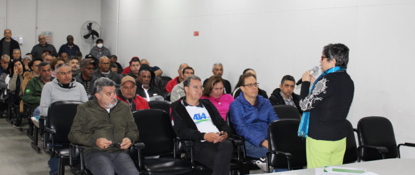 A imagem mostra uma sala de reuniões ou auditório com dezenas de pessoas sentadas, assistindo atentamente a uma mulher que fala ao microfone. A palestrante está de pé, de costas parcialmente voltadas para a câmera, e usa calça verde-limão, blusa preta com detalhes brancos nas mangas e um cachecol azul.  O público é composto majoritariamente por adultos e idosos, homens e mulheres, vestindo roupas casuais de frio. Algumas pessoas usam crachás e há cadeiras pretas organizadas em fileiras. O ambiente é simples, com paredes brancas e iluminação artificial. Ao fundo, um ventilador de parede está ligado e algumas pessoas permanecem em pé.  A cena sugere uma reunião comunitária ou capacitação, com foco em participação cidadã ou assuntos públicos.