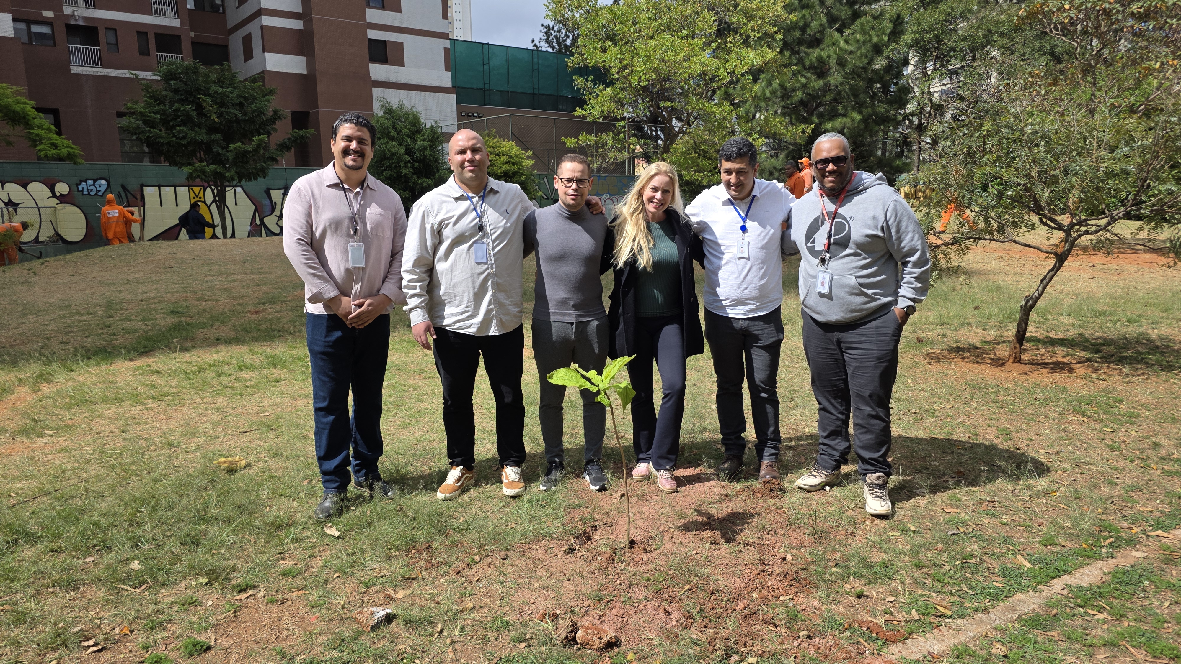 Imagem 1: Grupo de pessoas, três homens ao lado esquerdo, uma mulher de cabelos loiros ao centro e  mais dois homens ao lado direito, reunidos em área verde após o plantio de uma muda de árvore.  Imagem 2: Dois homens, um de camisa cinza e outro de camisa branca realizando o plantio de uma muda de árvore em área verde. 