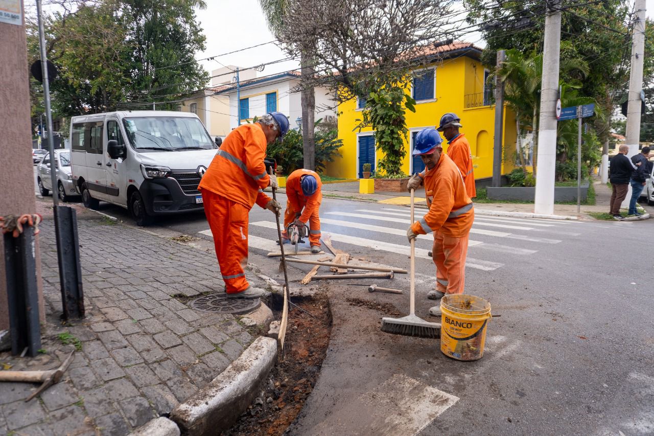Foto 1: Trabalhador com uniforme laranja, capacete branco e luvas verdes realiza manutenção em bueiro, utilizando uma barra de ferro para o serviço.  Foto 2: Linha de policiais militares perfilados ao lado de viaturas, com uma igreja amarela ao fundo em cenário urbano.  Foto 3: Grupo de profissionais da saúde com jalecos brancos sorriem em frente a uma tenda da Subprefeitura de Santo Amaro, com mesa contendo vacinas, caixa de descarte e materiais médicos.  Foto 4: Trabalhadores da prefeitura, com uniformes laranja e capacetes azuis, realizam manutenção no asfalto em uma esquina de bairro residencial.  Foto 5: Grupo de policiais militares fardados, em formação, conversa com um homem de camisa azul em uma rua movimentada.