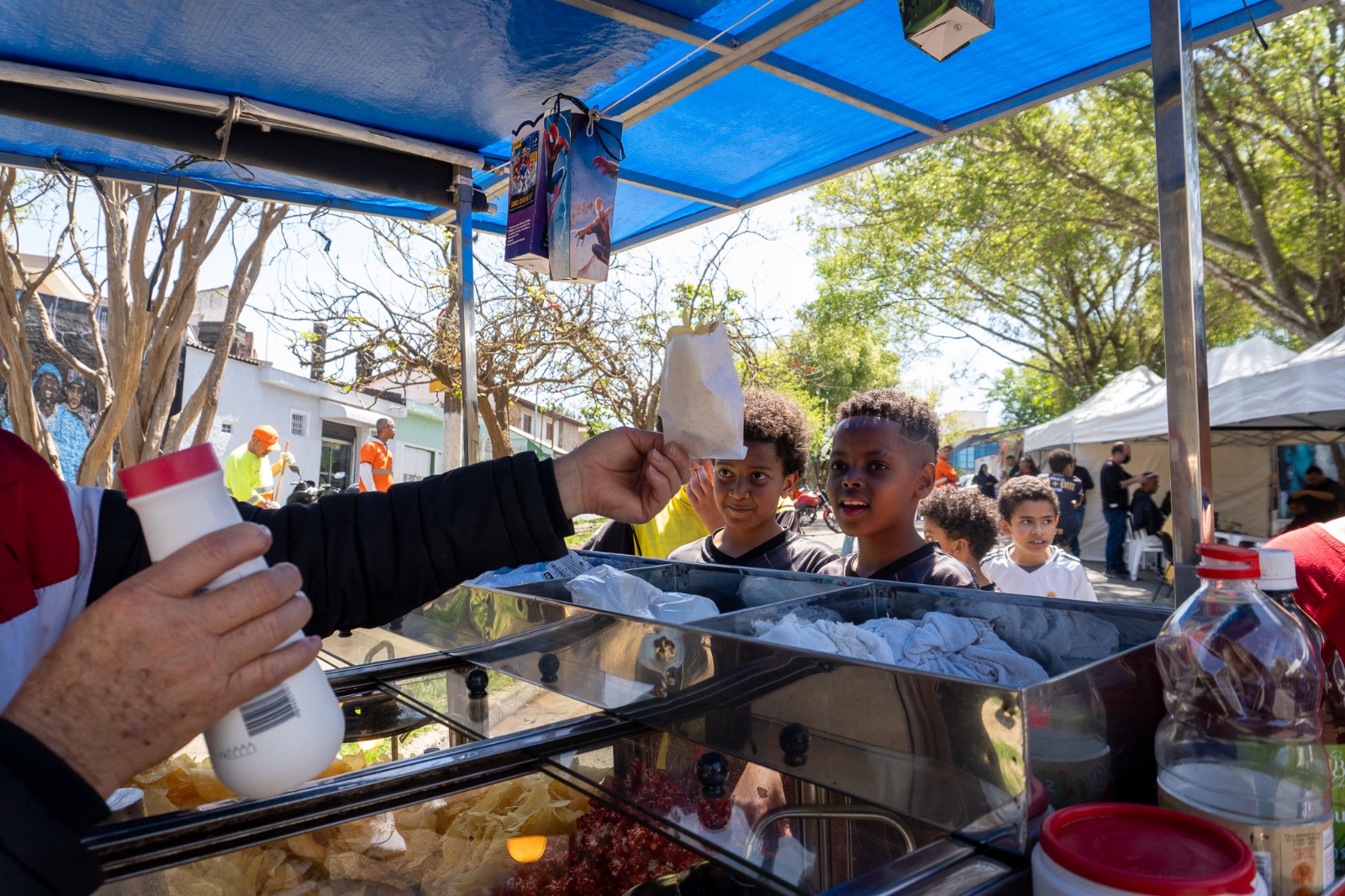 Primeira foto: Homem corta o cabelo de outro em ação comunitária ao ar livre, com o logo da subprefeitura de Santo Amaro ao fundo.  Segunda foto: Crianças aguardam animadas para receber saquinhos de pipoca em barraca durante evento de rua.