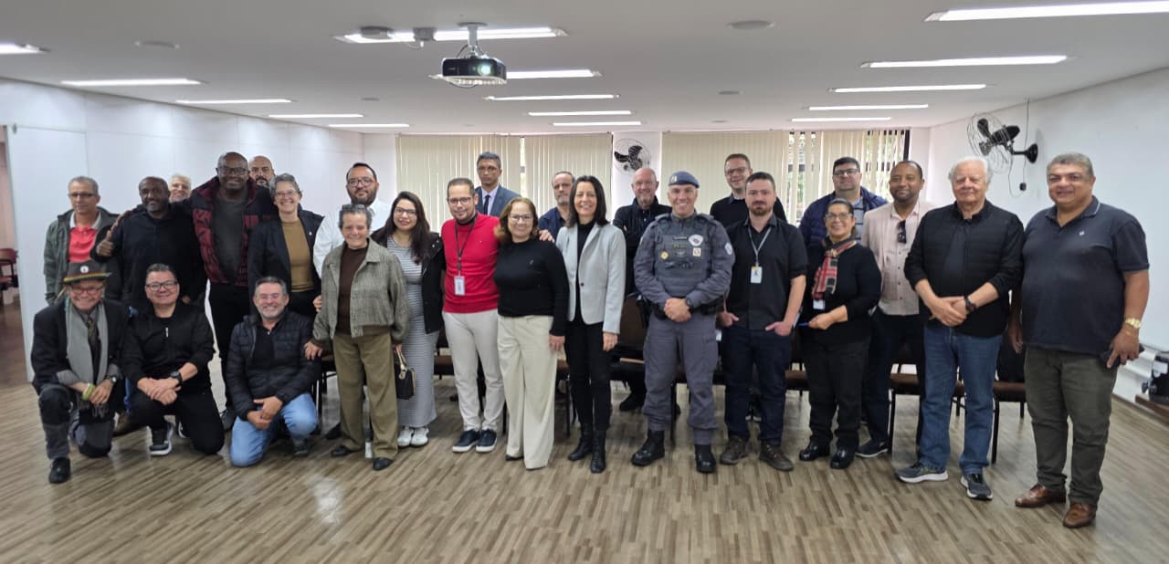 Descritivo das imagens:  Primeira imagem: grupo de pessoas posa sorrindo em uma sala de reuniões, entre elas servidores públicos, representantes da Polícia Militar e lideranças locais.  Segunda imagem: um policial fardado fala ao microfone durante reunião, enquanto outro homem com blusa vermelha e crachá o observa ao lado.