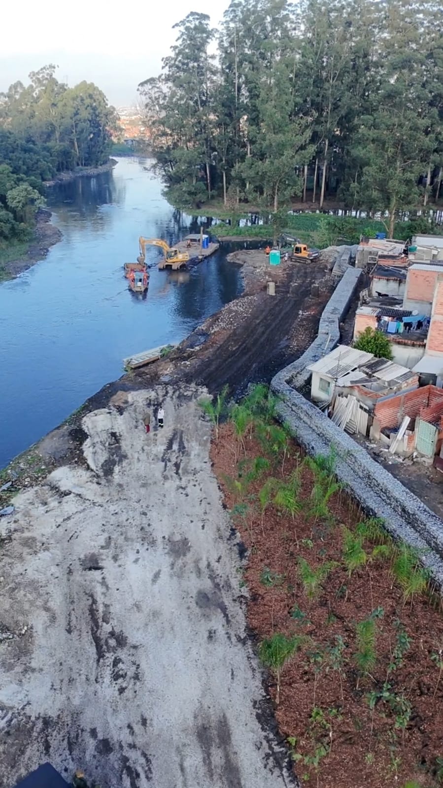 Foto aérea de um rio cercado por árvores e casas. Máquinas escavadeiras trabalham às margens da água em uma área de contenção com pedras e terra, enquanto uma faixa de vegetação recém-plantada aparece em primeiro plano. A imagem transmite uma grande obra de recuperação e proteção das margens do rio.