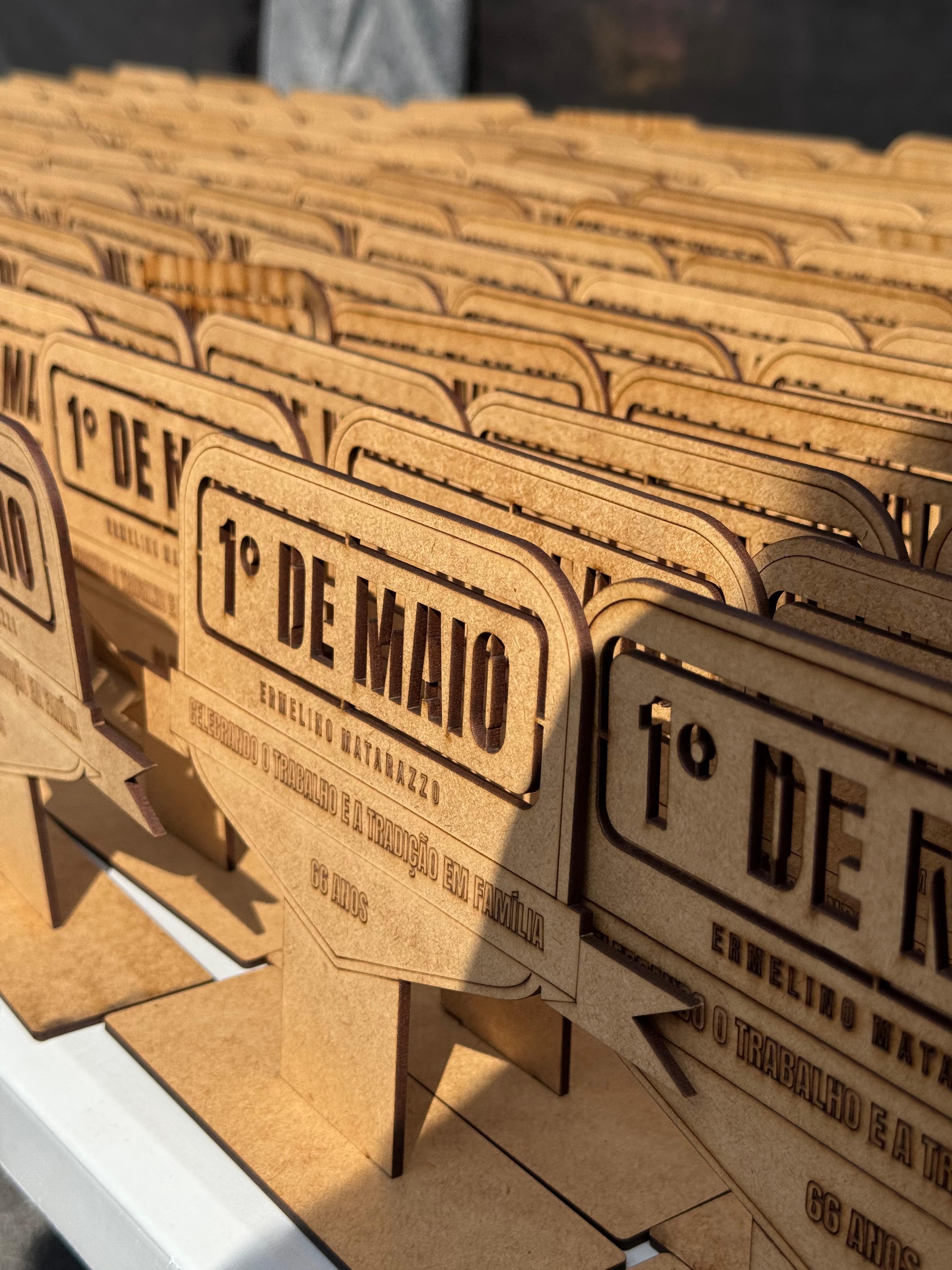  #praCegoVer  A imagem exibe uma série de objetos de madeira clara, possivelmente MDF, cortados a laser e gravados, dispostos em fileiras sobre uma superfície clara. Cada objeto tem um formato semelhante a um letreiro vintage, com as palavras "1º DE MAIO" em destaque na parte superior. Abaixo, em letras menores, lê-se "ERMELINO MATARAZZO" e, em seguida, a frase "CELEBRANDO O TRABALHO E A TRADIÇÃO EM FAMÍLIA", finalizando com "60 ANOS". As bordas dos objetos e as letras gravadas apresentam um tom mais escuro, resultado do processo de corte a laser. A profundidade de campo da foto destaca os objetos em primeiro plano, enquanto os demais se estendem em perspectiva, criando um efeito de repetição.