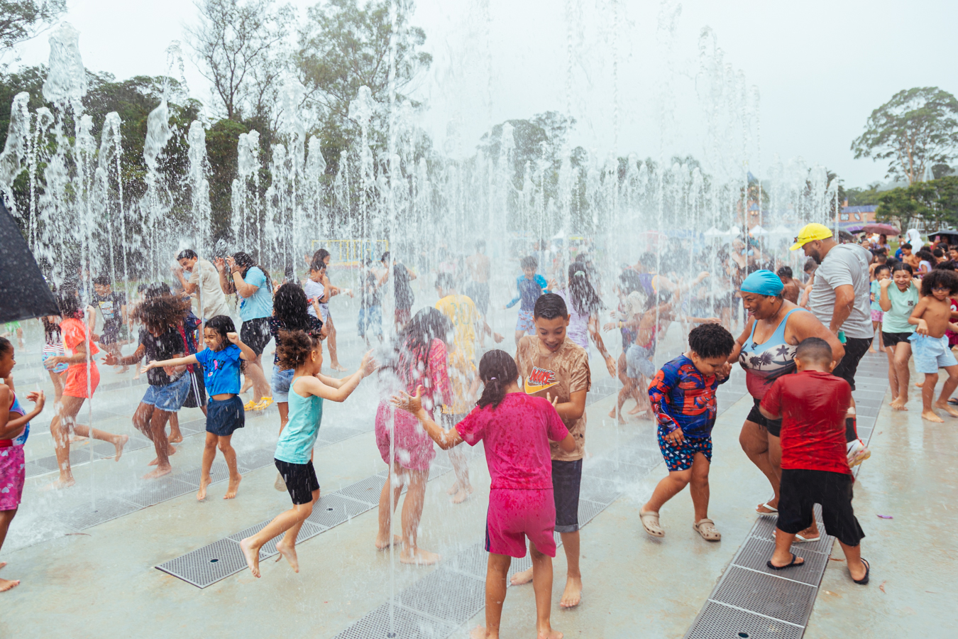 Imagem em ambiente externo, mostrando uma área de lazer com várias fontes de água jorrando para cima, formando colunas e cortinas de água transparente e esbranquiçada. O chão é claro, em tom de cinza bem claro, com grelhas metálicas retangulares distribuídas entre os jatos de água.  No centro e por toda a cena, há muitas crianças brincando e correndo entre as fontes. Elas estão descalças e usam roupas leves e coloridas, como camisetas e shorts em tons de rosa, vermelho, azul, verde, amarelo e laranja. Algumas roupas estão encharcadas e coladas ao corpo. As crianças aparecem rindo, pulando, esticando os braços para tocar a água e se protegendo dos jatos, criando uma sensação de movimento e alegria.  À direita da imagem, há alguns adultos acompanhando as crianças. Uma mulher usa um lenço azul na cabeça e uma blusa clara, enquanto um homem ao lado dela usa boné amarelo e camiseta clara. Ao fundo, é possível ver mais pessoas e barracas ou estruturas em cores claras, além de muitas árvores altas com copas verdes, que formam uma moldura natural para o espaço.  O céu está claro, em tom branco-acinzentado, e a luz é difusa. No conjunto, a imagem é muito colorida e vibrante, com o branco e o transparente da água contrastando com o colorido das roupas e o verde das árvores, transmitindo um clima de diversão, calor e brincadeira coletiva.