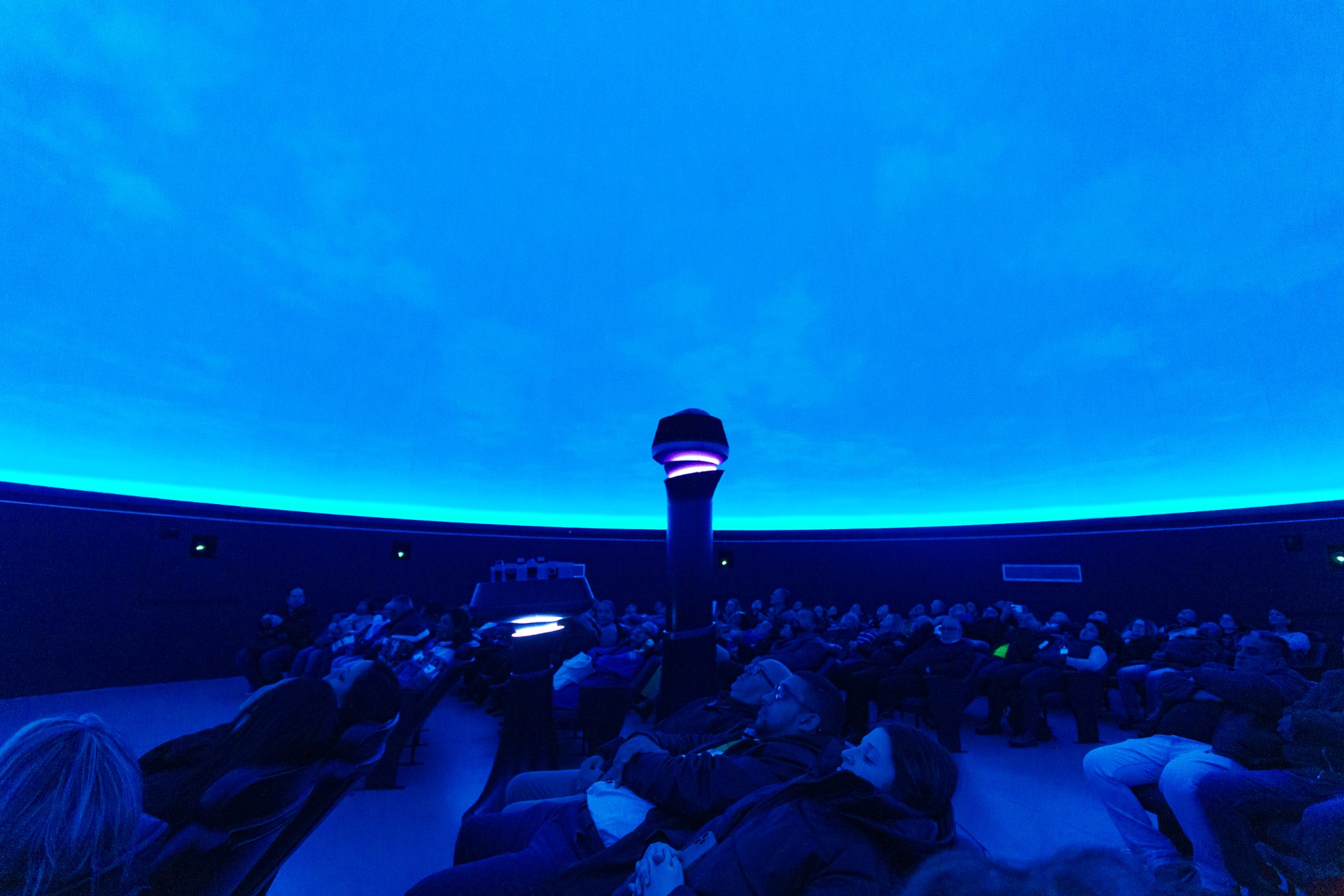 O interior de um planetário é mostrado durante uma sessão de projeção. O ambiente tem formato circular e é dominado por uma grande cúpula iluminada em diferentes tons de azul, que vão do azul profundo ao azul-turquesa brilhante próximo às bordas. No centro da sala está o projetor do planetário, uma estrutura cilíndrica preta, com detalhes iluminados em roxo-violeta e branco. As paredes escuras, quase pretas, contornam o espaço e apresentam pequenos pontos de luz verde, reforçando a atmosfera tecnológica e imersiva.  Ao redor do projetor, dezenas de pessoas estão sentadas em poltronas reclinadas, observando a projeção no teto. A iluminação azul intensa faz com que os rostos e as roupas apareçam pouco definidos, predominando tons de preto, cinza e azul-escuro. Em primeiro plano, algumas pessoas podem ser vistas mais claramente, reclinadas e olhando para cima. A cena transmite uma sensação de tranquilidade, contemplação e encantamento, típica de uma experiência de observação do céu em um planetário.