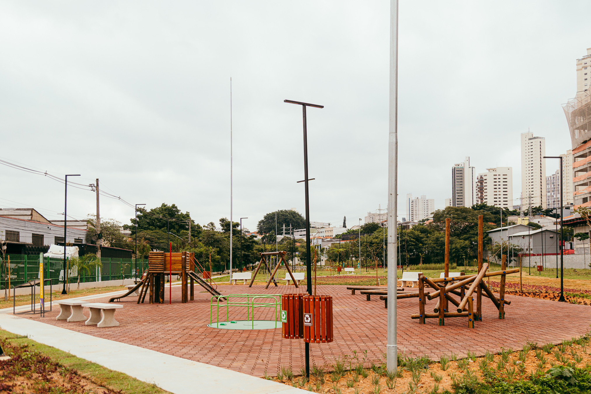 Fotografia de uma área de lazer do recém-inaugurado Parque Verde da Mooca. Em primeiro plano, há um playground com brinquedos de madeira, incluindo escorregador, balanços e estruturas para escalada, instalados sobre piso intertravado avermelhado. Bancos de concreto e lixeiras estão distribuídos pelo espaço. Ao fundo, extensas áreas gramadas com árvores jovens e postes de iluminação acompanham os caminhos do parque. No horizonte, prédios residenciais e comerciais contrastam com a vegetação, evidenciando a integração do novo parque ao ambiente urbano da Mooca. O céu está nublado.