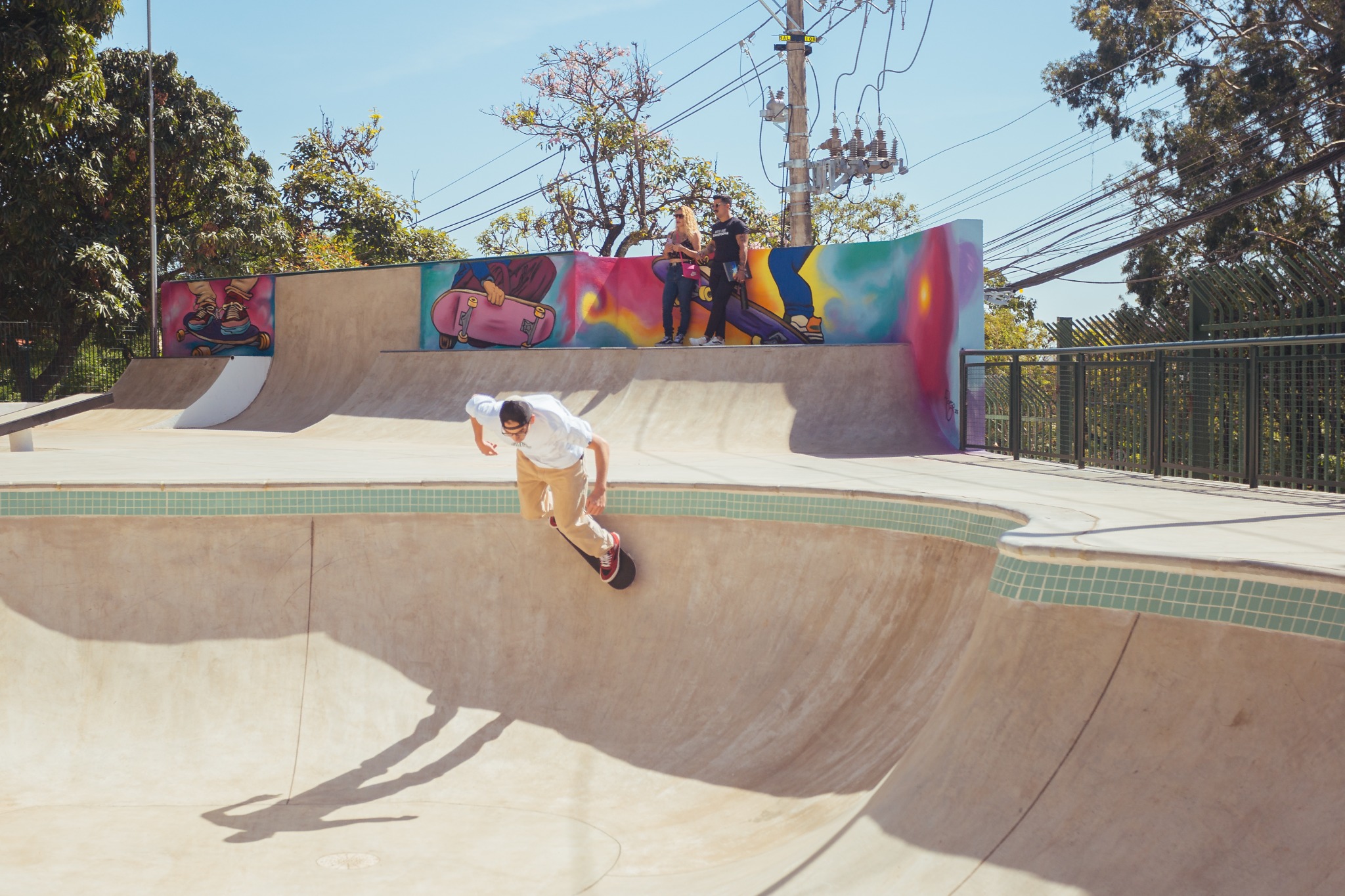 Imagem mostra uma pista de skate ao ar livre em dia ensolarado. Em primeiro plano, um jovem anda de skate dentro de uma pista em formato de bowl, inclinando o corpo enquanto desliza pela parede curva de concreto. A sombra do skatista se projeta na superfície da pista.  Ao fundo, há uma rampa vertical com mural colorido que retrata figuras e elementos ligados ao universo do skate. Duas pessoas observam a cena sobre a plataforma da rampa. O espaço é cercado por grades metálicas e árvores, com postes e fiação elétrica visíveis ao redor, indicando ambiente urbano integrado a área verde.