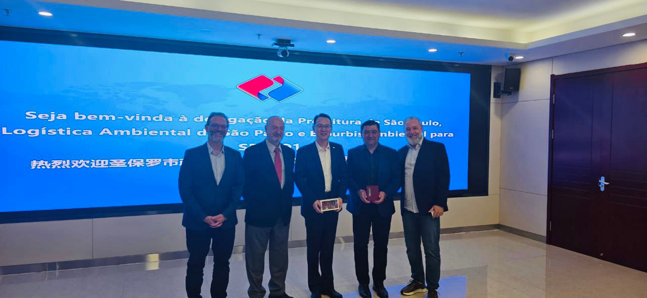 Quatro homens posam sorrindo diante de uma tela azul com mensagens de boas-vindas em português e chinês, em um ambiente moderno e iluminado.