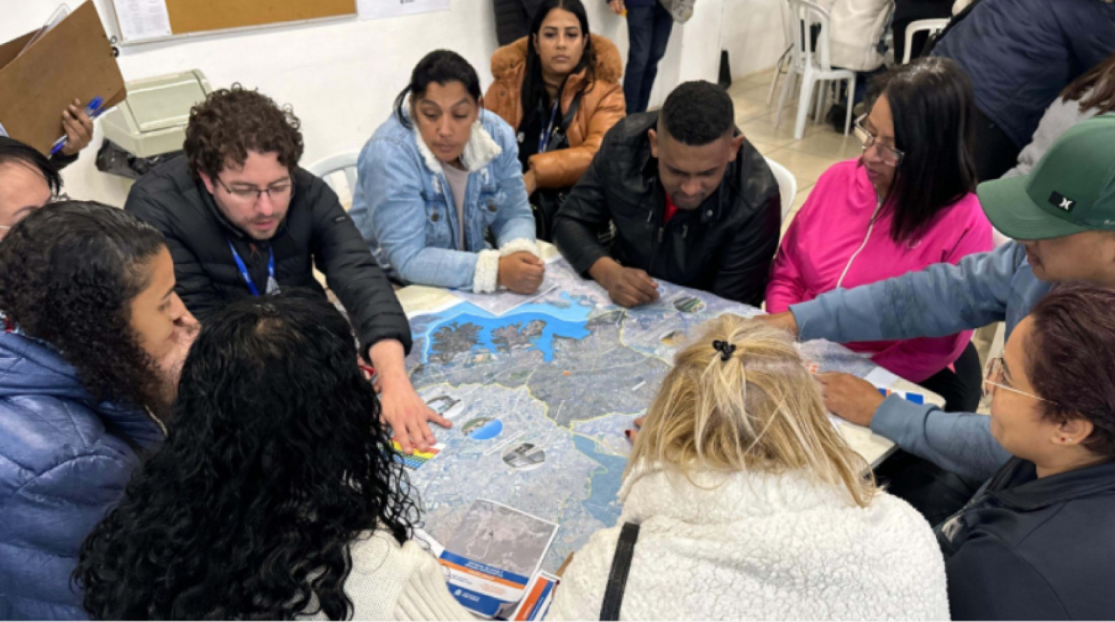A imagem mostra um grupo de pessoas sentadas ao redor de uma mesa, participando de uma atividade coletiva. Elas estão reunidas em um ambiente interno, parecido com uma sala de reunião ou centro comunitário. Sobre a mesa há um grande mapa colorido espalhado, e os participantes apontam e discutem diferentes pontos nele. O grupo é diverso, composto por homens e mulheres de idades variadas, todos atentos e envolvidos na atividade. Algumas pessoas usam casacos, sugerindo que o clima está frio. Ao fundo, há cadeiras e quadros na parede, indicando um espaço simples e funcional.