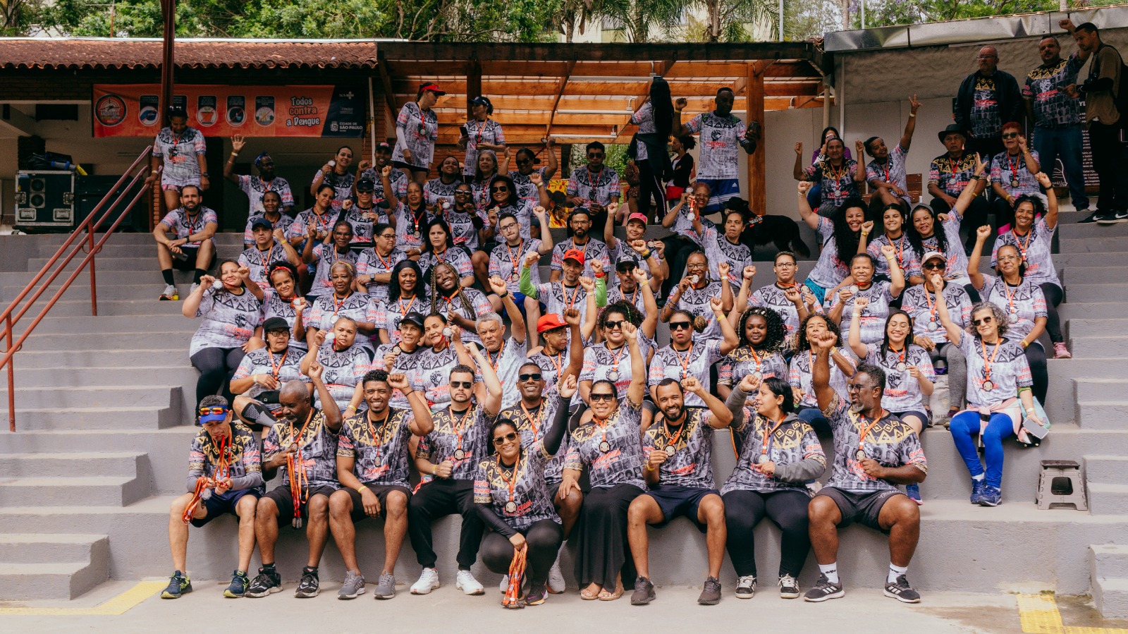 Foto de um grande grupo de pessoas reunidas em arquibancadas ao ar livre, todas usando camisetas coloridas de padrão geométrico, com medalhas no pescoço. Elas posam sorrindo, levantando os braços ou fazendo gestos de comemoração. Há diversidade de idade, gênero, cor e estilos entre os participantes. Ao fundo, vê-se uma estrutura de madeira e árvores. O clima é de celebração, união e conquista coletiva.