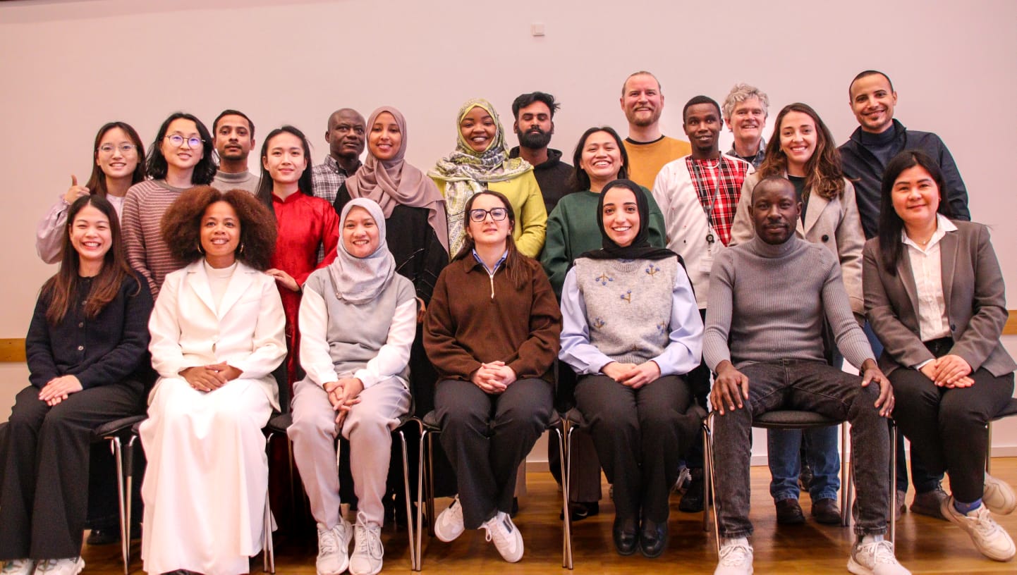 Grupo de cerca de 20 pessoas posando em duas fileiras (sentadas e em pé) em uma sala clara. São diversas em aparência e vestimenta, com roupas formais e casuais. A maioria sorri para a câmera, transmitindo um clima de diversidade e acolhimento.