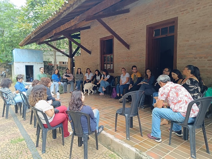 Grupo de pessoas sentadas em círculo, em cadeiras, participando de uma conversa ao ar livre, na varanda de uma casa simples com paredes de tijolo e telhado de madeira. Há árvores ao redor e um cachorro no centro do grupo.