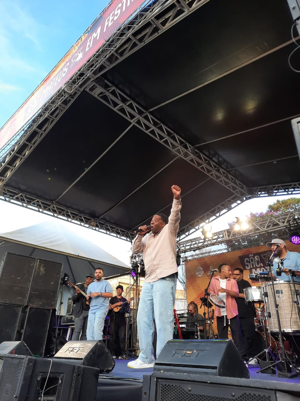 #PraTodosVerem: O cantor Netinho de Paula no centro do palco, vestindo camisa bege e calça jeans clara, segura o microfone e levanta o braço direito em gesto de celebração. Ao fundo, músicos tocam cavaquinho, pandeiro e outros instrumentos.