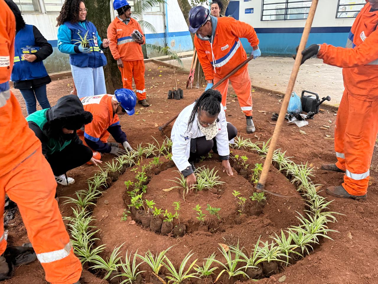 #PraTodosVerem: Quatro trabalhadores de uniforme laranja trabalham cavando a terra e organizando as mudas plantadas. Uma mulher de uniforme branco ajuda, e de fundo outros dois homens de uniforme laranja conversam com pessoas.