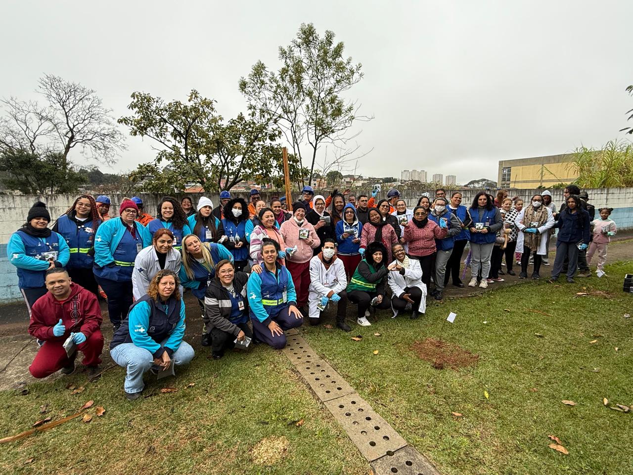 #PraTodosVerem: Grupo grande de pessoas unidas posando para foto. Ambiente ao ar livre, com grama e algumas árvores de fundo. É dia e está nublado.