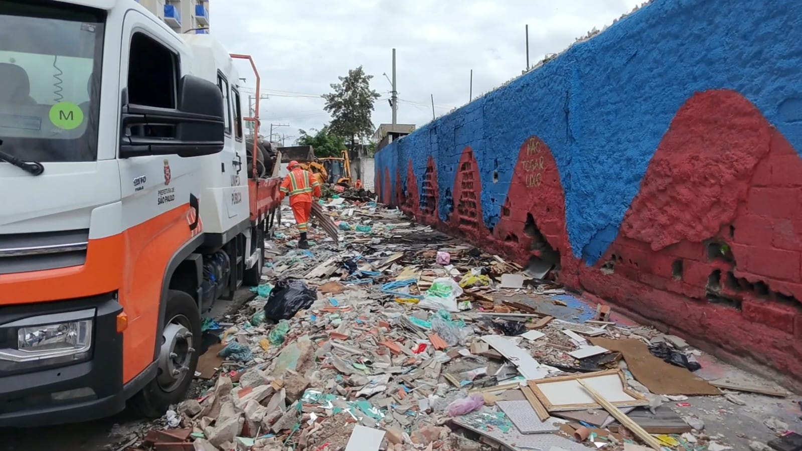 Imagem mostra uma ação de zeladoria urbana: caminhão da Prefeitura de São Paulo e trabalhadores realizando a retirada de grande quantidade de lixo e entulho espalhados ao longo de uma via. À direita, há um muro pintado de azul e vermelho, enquanto o chão está tomado por resíduos de construção e descarte irregular.
