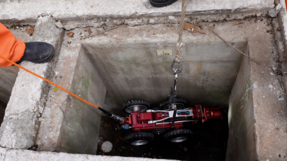 A imagem, capturada de um ângulo superior, mostra o interior de um bueiro de concreto de formato quadrado. No fundo da cavidade, encontra-se um pequeno robô de inspeção remoto na cor vermelha metálica, equipado com quatro rodas pretas de borracha tratoradas. O robô está conectado a um cabo laranja fino que se estende para fora do bueiro, em direção ao canto superior esquerdo, onde se vê parte da calça laranja e a bota preta de um operador. O robô também está preso a uma corda de sustentação por um mosquetão metálico, utilizada para descê-lo ou içá-lo. As paredes internas do bueiro são de concreto cinza e o fundo apresenta um pouco de terra e pequenos detritos.