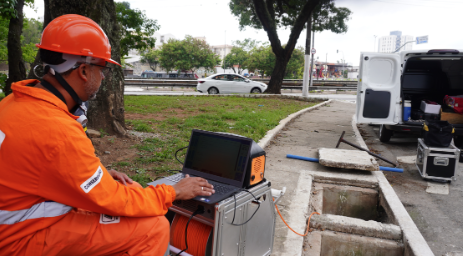 A imagem mostra um homem negro, de perfil, trabalhando ao ar livre em uma calçada. Ele veste um uniforme de trabalho laranja vibrante com faixas refletivas cinzas e usa um capacete de segurança também laranja. O homem está agachado à esquerda, operando um notebook que está apoiado sobre uma maleta técnica prateada. À sua frente, há uma tampa de bueiro aberta no chão, revelando uma cavidade quadrada de concreto. Um cabo laranja sai do equipamento e desce para dentro do bueiro, sugerindo uma inspeção de tubulação ou rede subterrânea. Ao fundo, vê-se um gramado com árvores, uma rua com um carro branco passando e uma van branca estacionada com as portas traseiras abertas, revelando mais ferramentas e equipamentos. O dia parece nublado e o ambiente é urbano.