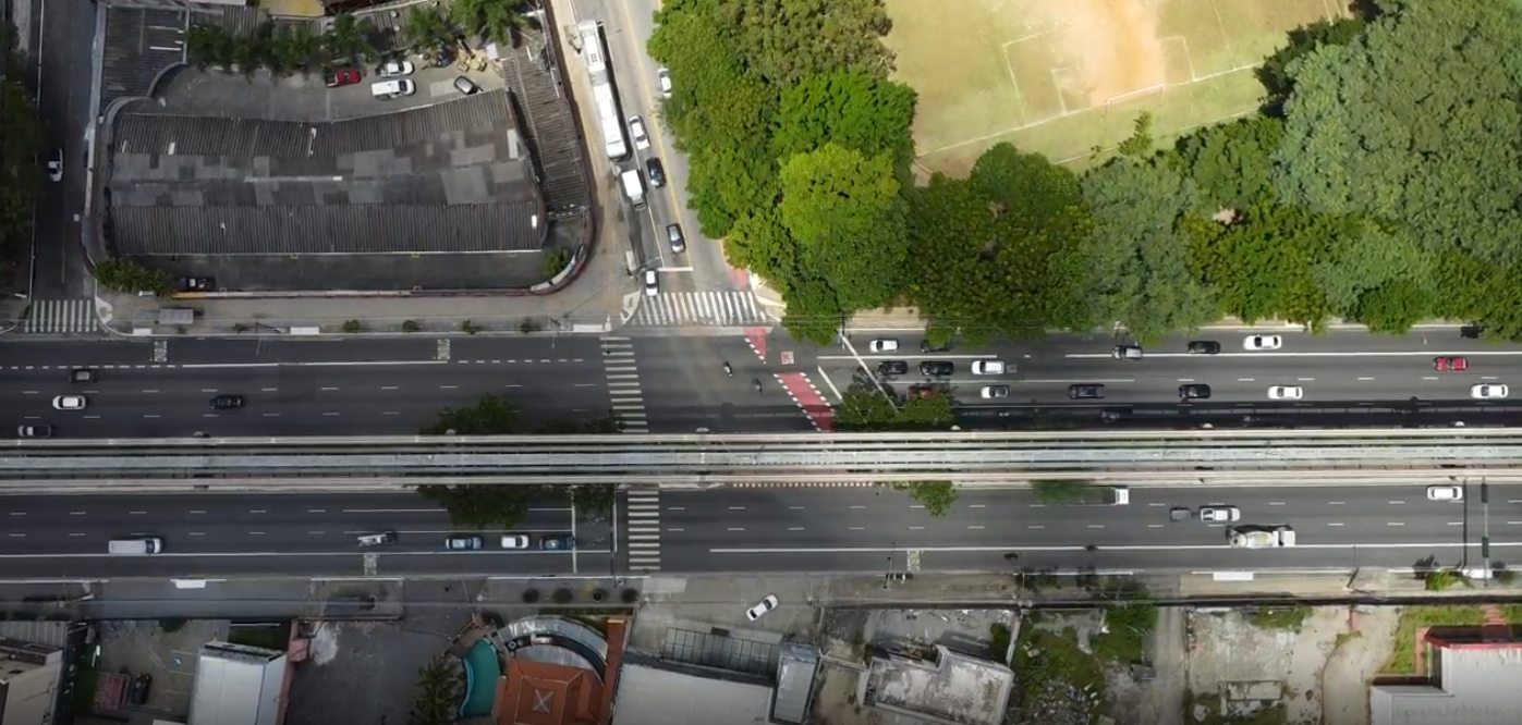 A imagem é uma fotografia aérea em plano aberto, tomada verticalmente de cima para baixo, mostrando um trecho de uma larga avenida urbana. O centro da imagem é cortado horizontalmente pelos trilhos de concreto de um monotrilho elevado. Abaixo da estrutura do monotrilho, a avenida possui asfalto escuro com sinalização de faixas brancas nítidas, indicando recapeamento recente, com carros, caminhões e ônibus transitando em ambos os sentidos. À esquerda, há um cruzamento com uma rua perpendicular, marcado por faixas de pedestres zebradas. A parte superior da foto exibe uma área com muitas árvores verdes densas, parte de um campo de futebol gramado e o telhado de um grande galpão comercial. A parte inferior mostra uma mistura de telhados residenciais e comerciais, pátios de concreto e uma pequena piscina azul em uma propriedade privada.